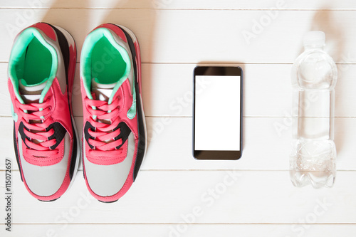 Red sneakers with bottle with water, smartphone with copy space on screen on white wooden background indoors. Smartphone mockup. Sports and fitness background, healthy lifestyle concept.