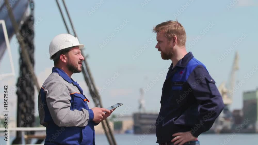 Two skilled workers at a port with a tablet PC, nearby cargo ship partly visible Two skilled workers at a port with a tablet PC, nearby cargo ship partly visible