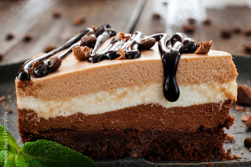 piece of layered cake with chocolate topping close-up on black plate, blurred wooden background