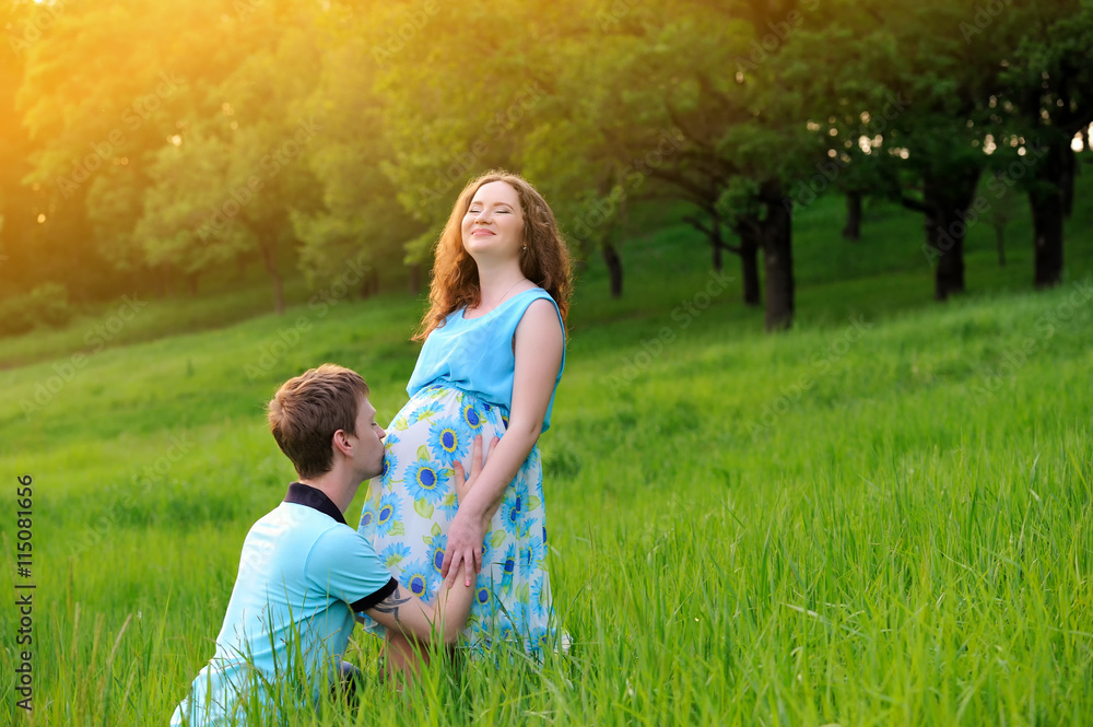 Fototapeta premium Happy pregnant couple standing on meadow at sunset. The guy kisses the girl belly. The girl laughs with delight. Portrait. Horizontal orientation