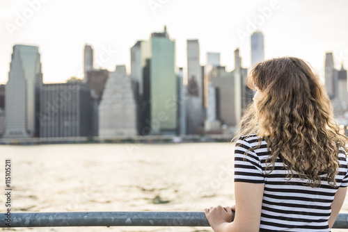 Young woman in striped shirt looking at Manhattan skyline at Brooklyn bridge park during sunset
