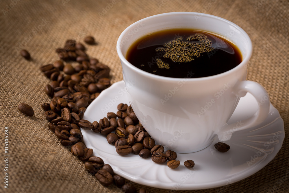 Tasse Kaffee mit Untertasse und Kaffeebohnen auf Leinentuch, Nah Tasse Kaffee mit Untertasse und Kaffeebohnen auf Leinentuch, Nah