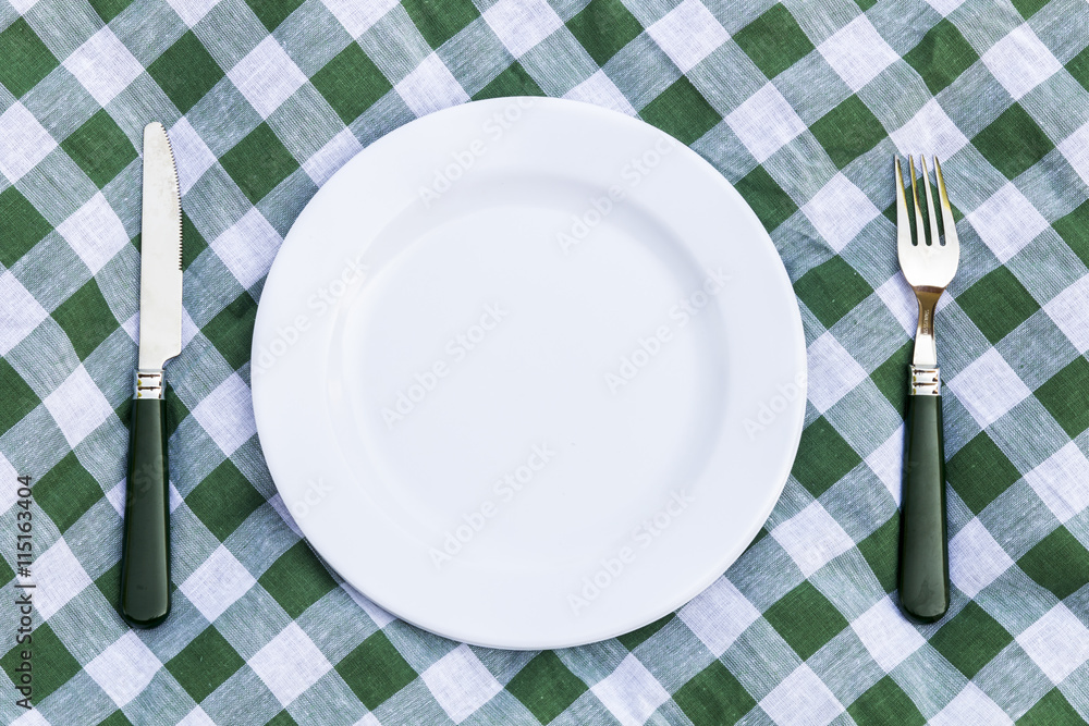 Knife, white plate and fork on green picnic tablecloth. Flat lay Knife, white plate and fork on green picnic tablecloth. Flat lay