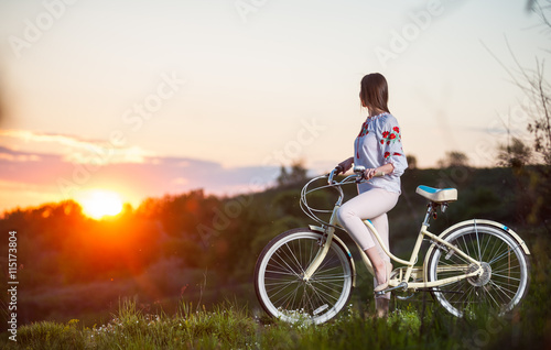 Beautiful sunset and girl in Ukrainian embroidery with retro bike enjoying them. Blurred background