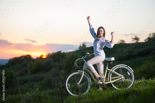 Smiling woman in Ukrainian embroidery with retro bike standing at hill and showing thumb up gesture of good class against beautiful sunset with a blurred background of greenery