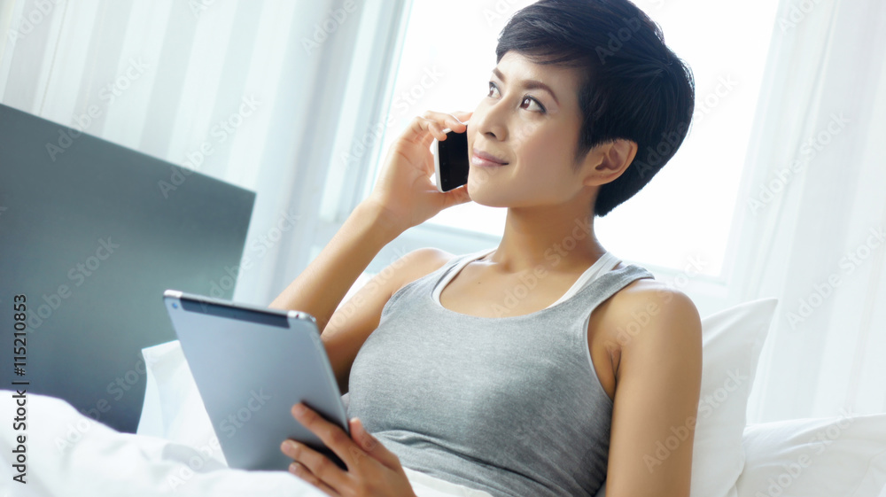 Asian Woman smiling and using tablet and cellphone, sitting in bed Asian Woman smiling and using tablet and cellphone, sitting in bed