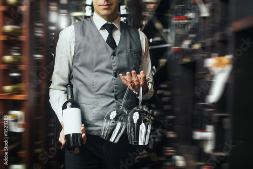 Close up of arms of skillful male sommelier standing in liquor store. He is holding a bottle of wine and two glasses. The man is smiling. Copy space in right side