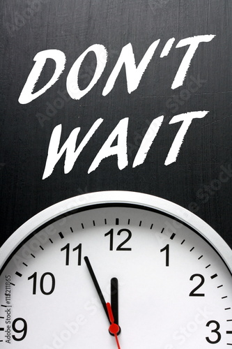 The words Don't Wait in white text on a blackboard above a modern wall clock with the time at almost midnight