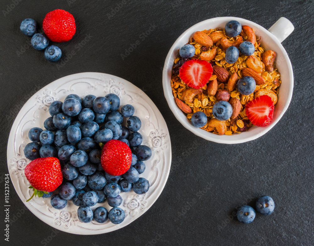 Breakfast bowl with granola made from oat flakes, dried fruits and nuts, and fresh blueberries and strawberries. Black stone background, top view. Breakfast bowl with granola made from oat flakes, dried fruits and nuts, and fresh blueberries and strawberries. Black stone background, top view.