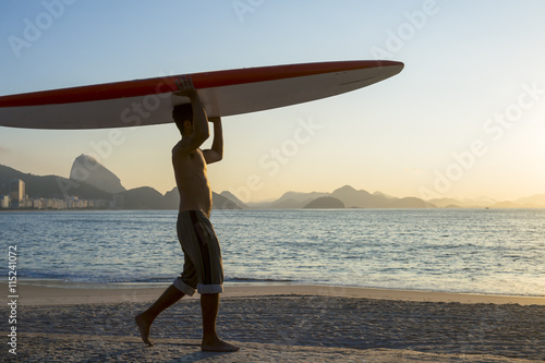 A young Brazilian man on Copacabana Beach balancing a surfboard on his head in front of a sunrise skyline view of Sugarloaf Mountain in Rio de Janeiro, Brazil