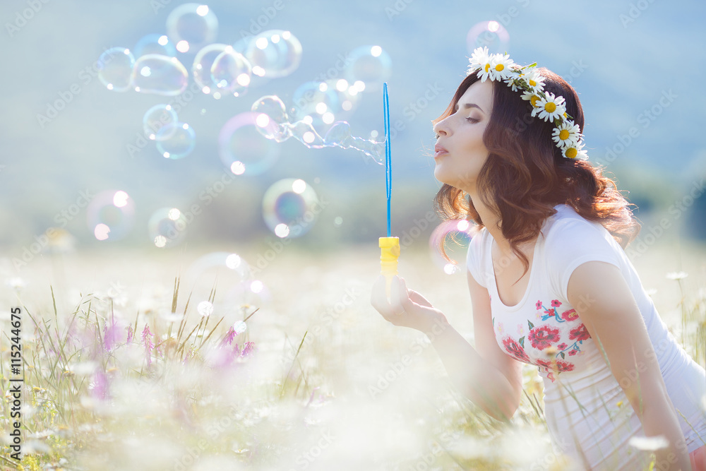 Beautiful pregnant woman with long curly brown hair, dressed in a white t-shirt without sleeves, on his head wearing a wreath of white daisies,blow bubbles on a white flowered field