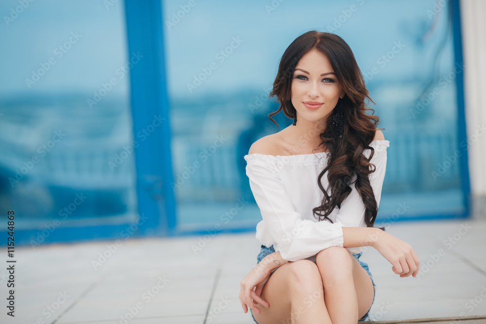 Young beautiful brunette woman with long curly hair , dressed in a white shirt and blue denim shorts,sitting in the open air in the summer on a background of show-windows with blue glass