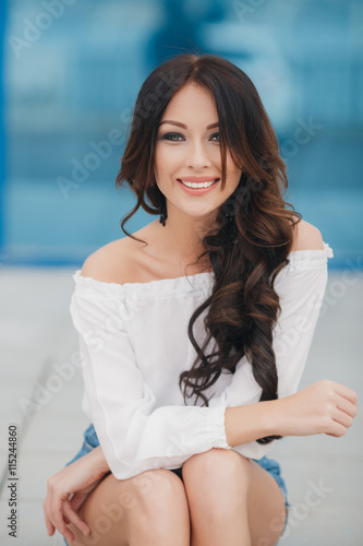 Free Download Young beautiful brunette woman with long curly hair , dressed in a white shirt and blue denim shorts,sitting in the open air in the summer on a background of show-windows with blue glass - GFXLoad