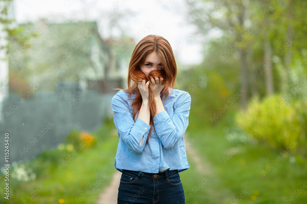Beautiful healthy young redhead woman hides her face in her hair posing on the green grass of country road Beautiful healthy young redhead woman hides her face in her hair posing on the green grass of country road