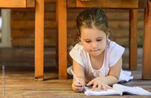 Lovely little girl lying down on the floor drawing on notebook outdoor on backyard terrace.  Childhood happiness concept.