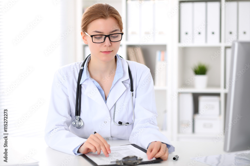 Young brunette female doctor sitting at the table and working at hospital office. Health care, insurance and help concept. Physician ready to examine patient Young brunette female doctor sitting at the table and working at hospital office. Health care, insurance and help concept. Physician ready to examine patient