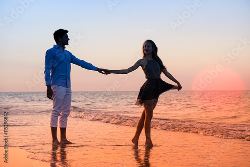 Silhouette of young couple walking at sunset on the beach - Happy lovers holding hands at night by the ocean with big red sun background - Iconic concept of love and tropical holidays with warm colors