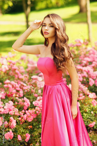 Young beautiful pretty woman posing in long evening luxury dress against bushes with pink roses on a sunny summer day. Vogue style fashion sensual portrait