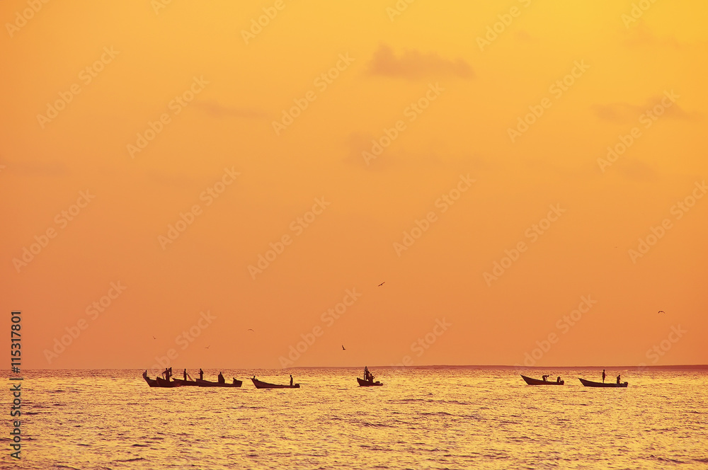 Fototapeta premium silhouettes of fishermen on the boats at sea. Orange sunset lighting. Birds in the sky circling above the boats 