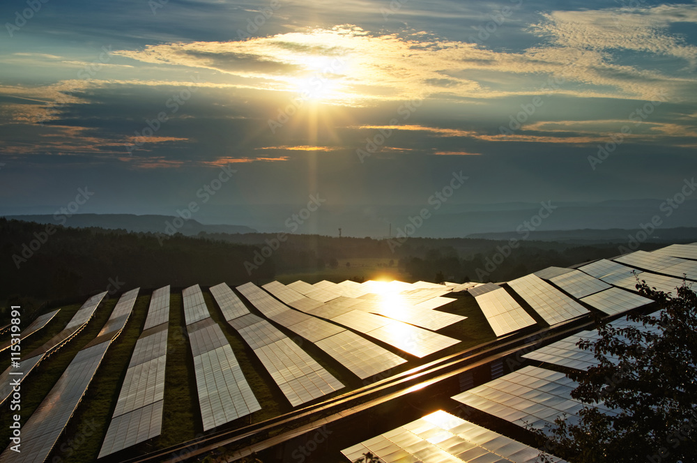 Solar power plant between forests at sunset with a star shining rays. The smoking factory chimneys and forested mountains fading into the mist in the distance.