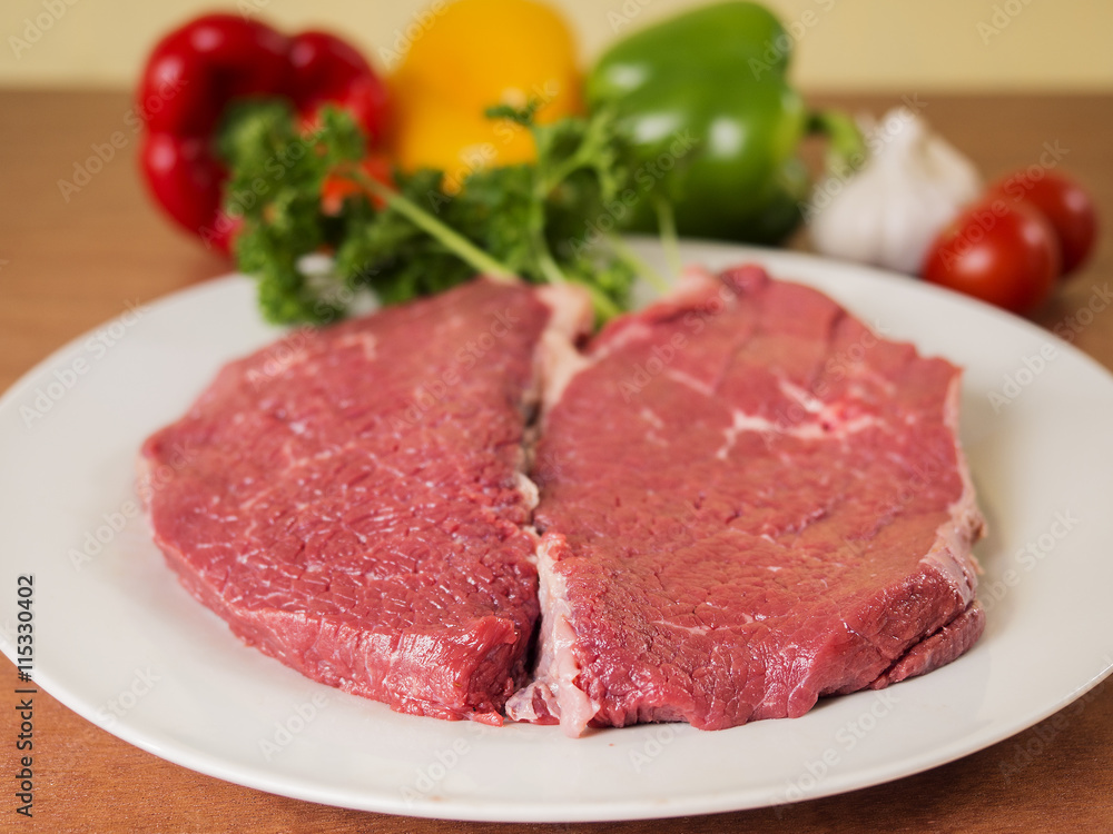 Two pieces of raw beef on a plate and some vegetables in background.