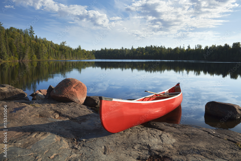 Red canoe on rocky shore of calm lake with pine trees - obrazy, fototapety, plakaty 