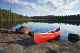 Red canoe on rocky shore of calm lake with pine trees