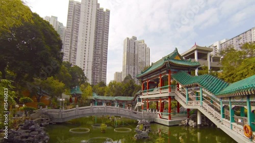 Urban residential towers with contemporary, highrise architecture overshadow a tranquil Buddhist temple garden with a pond and decorative fountain in Southeast Asia.