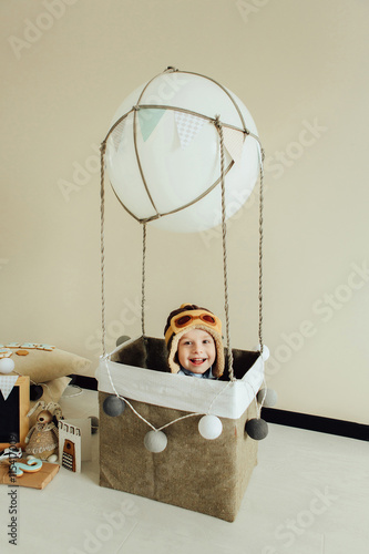 Happy kid playing in handmade basket of air balloon. Child having fun at home. Young pilot indoors at solid color background with copy space. Boy in hat looking at camera. Blank chalkboard.