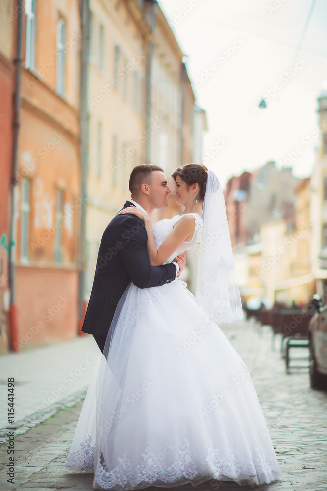 Naklejka premium Wedding photo shooting. Bride and bridegroom walking in the city. Married couple embracing and looking at each other. Holding bouquet. Outdoor, full body