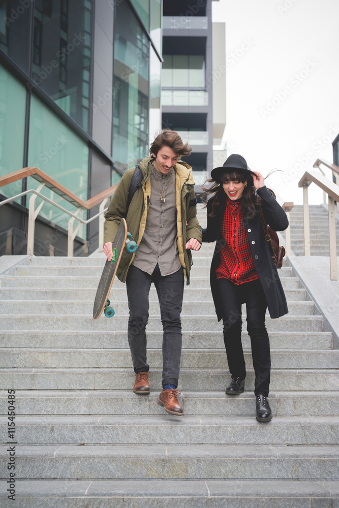 Obraz premium Young couple of caucasian woman and man, with skate and moustache, walking down the stairs hugging and smiling - youth, carefree, freshness concept