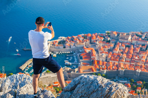 Male traveller make a panoramic photo of the old city of Dubrovnik from the mountain to his phone for Instagram or other social networks. Travel to Croatia. Summer vacation. © LALSSTOCK