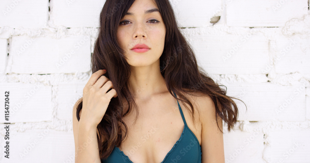 Seductive young woman with a sultry expression standing in front of a white brick wall looking at the camera with her hand to her long dark hair. Seductive young woman with a sultry expression standing in front of a white brick wall looking at the camera with her hand to her long dark hair.