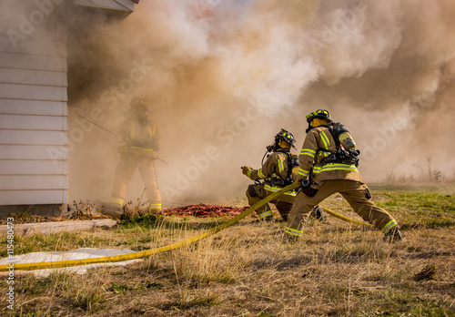 Fire fighters stand by with a hose while another fire fighter battles through heavy smoke to break open a window to allow water to be sprayed on the fire inside. 