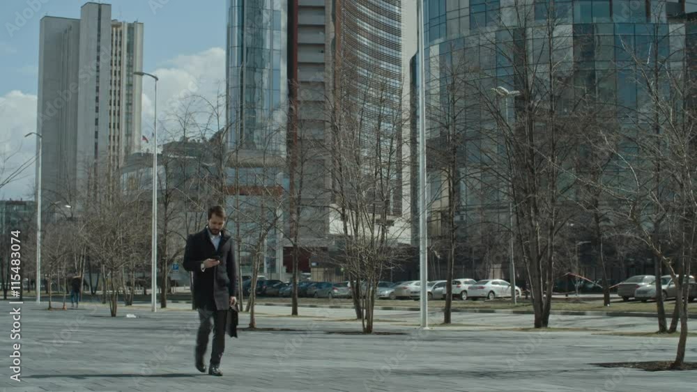 Businessman using his smart phone and answering a call while walking on the city street; modern glass office buildings in the background Businessman using his smart phone and answering a call while walking on the city street; modern glass office buildings in the background