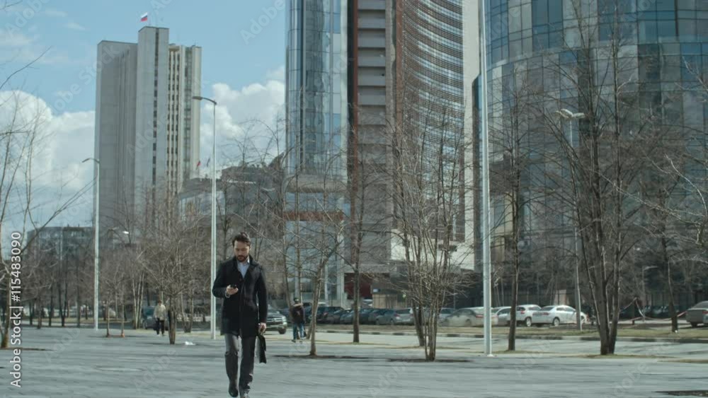Businessman with briefcase walking on the street and using his smart phone, then checking time on wristwatch and waiting for somebody Businessman with briefcase walking on the street and using his smart phone, then checking time on wristwatch and waiting for somebody