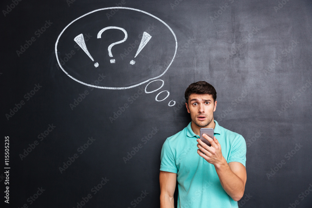 Amazed puzzled young man using cell phone over chalkboard background