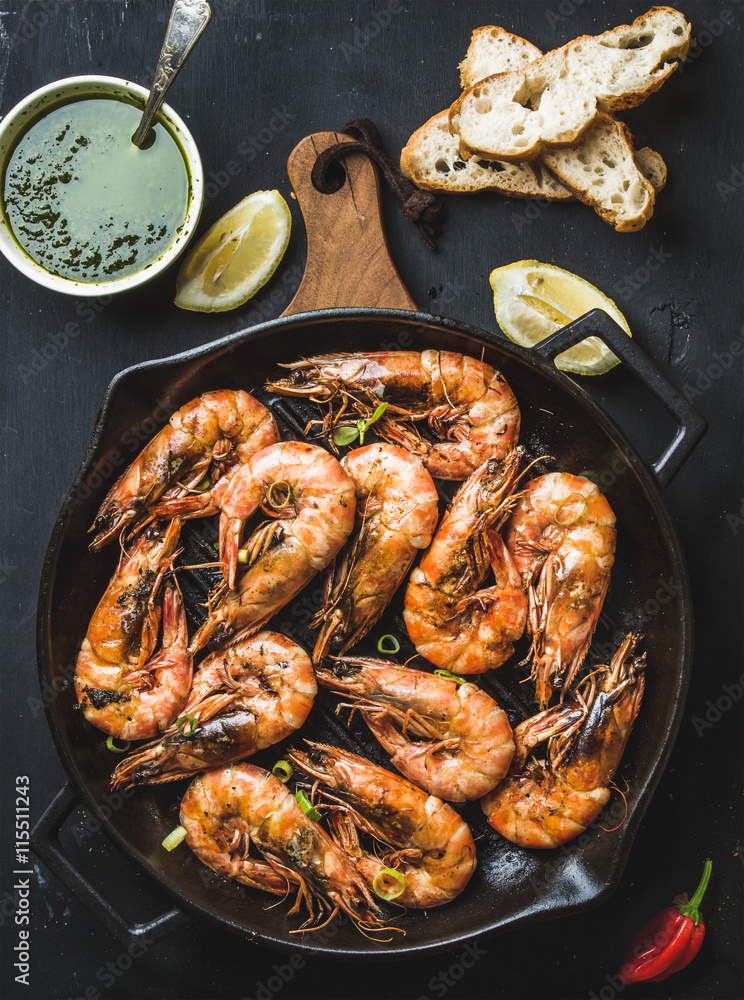 Roasted tiger prawns in iron grilling pan on wooden board with fresh leek, lemon slices, pepper, bread and pesto sauce over black background Roasted tiger prawns in iron grilling pan on wooden board with fresh leek, lemon slices, pepper, bread and pesto sauce over black background