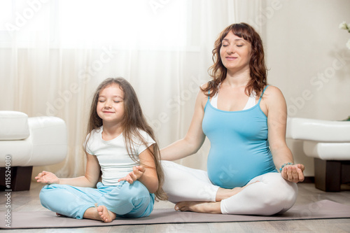 Family healthy lifestyle concept. Pregnancy Yoga and Fitness. Young happy pregnant yoga mom resting after workout with kid girl in living room. Pregnant mother and child meditating together at home