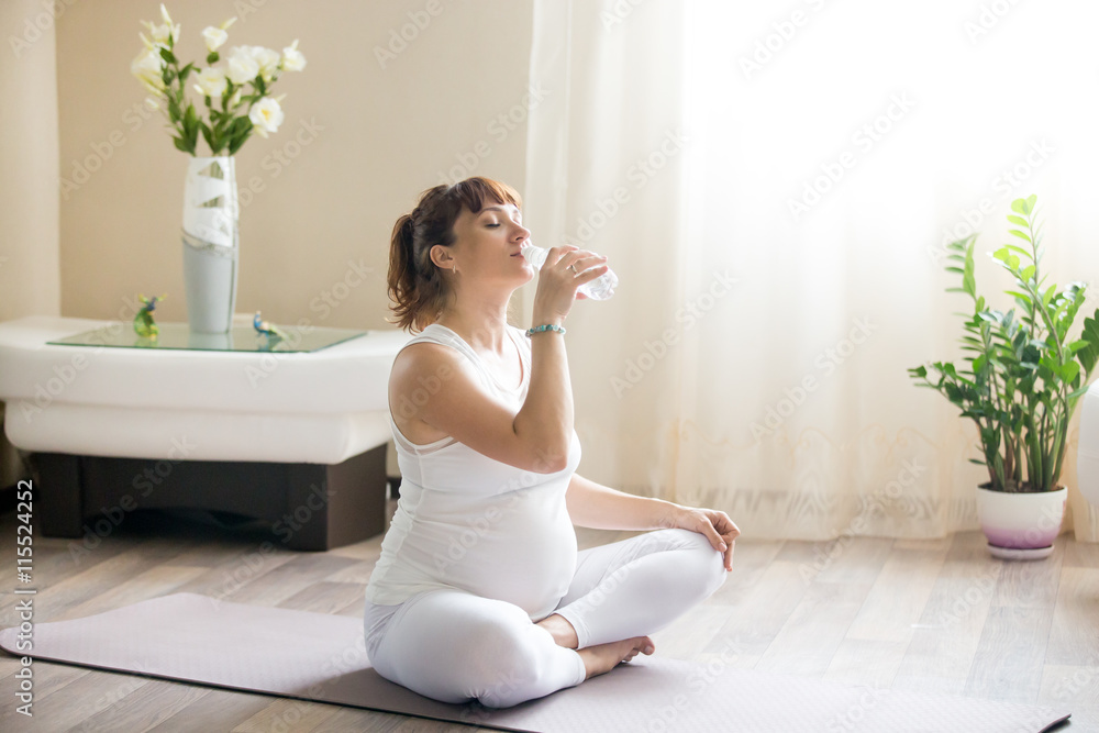 Portrait of young pregnant model drinking fresh water with closed eyes after working out in living room interior. Future mom expecting baby holding bottle indoors. Healthy maternity concept Portrait of young pregnant model drinking fresh water with closed eyes after working out in living room interior. Future mom expecting baby holding bottle indoors. Healthy maternity concept