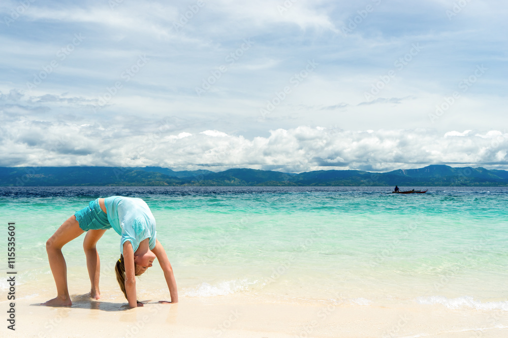 young slim teen girl do gymnastic exercise at white sand beach of tropical sea under blue sky