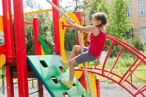 Little caucasian girl dressed in a red jersey playing on playground, climbing the wall on a rope