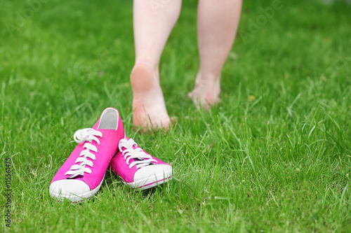 Female sneakers laying on green grass with female barefoot legs on background