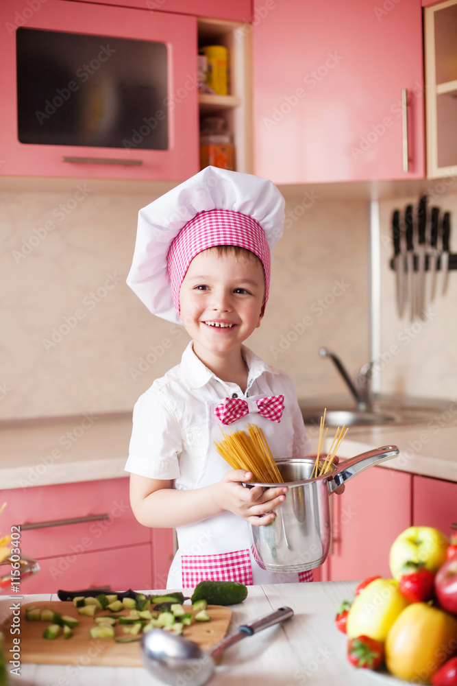 portrait of little boy in the hat of the chef and an apron. Little cooks chef in the kitchen preparing spaghetti. Emotional pictures portrait of little boy in the hat of the chef and an apron. Little cooks chef in the kitchen preparing spaghetti. Emotional pictures