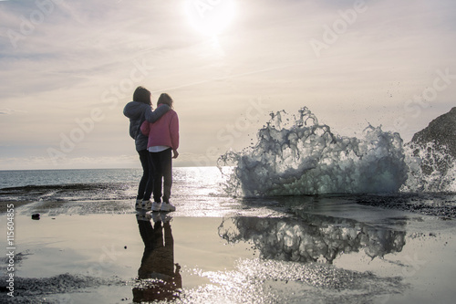 Amiche abbracciate al mare al tramonto che guardano un'onda antropomorfa minacciosa. La situazione è riflessa su  uno specchio d'acqua.  photo