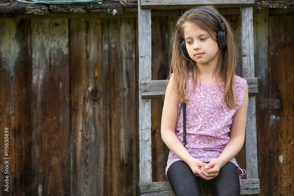 Little sad girl with headphones sitting on a wooden stepladder in the village outdoors. Little sad girl with headphones sitting on a wooden stepladder in the village outdoors.