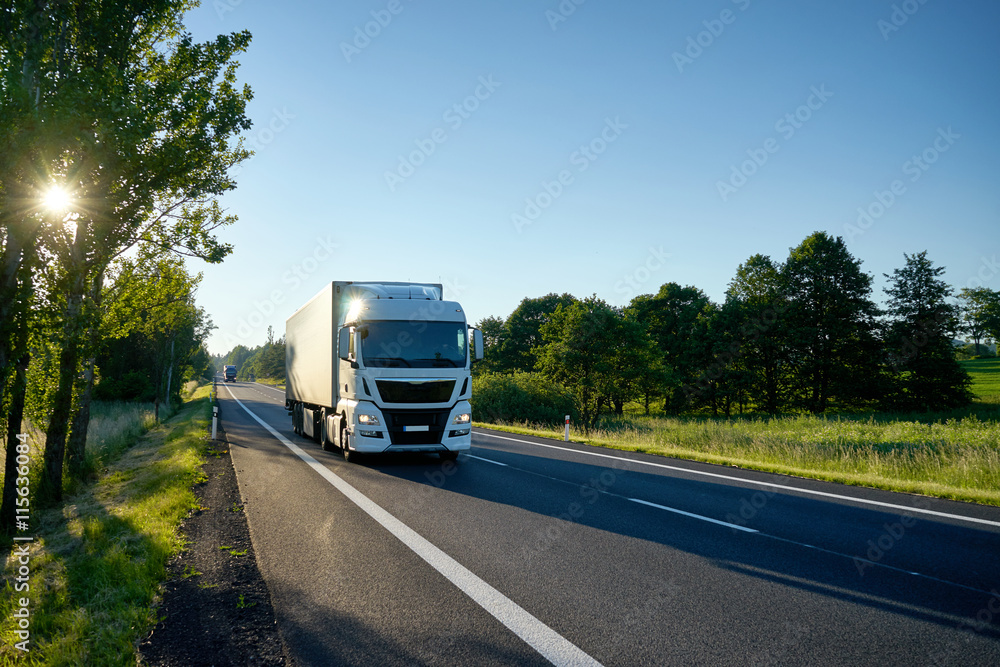 White truck arriving on the asphalt road. Stellar rays of the sun shining between deciduous trees. From a distance, second truck coming.