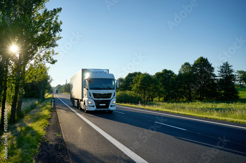 Obraz na płótnie White truck arriving on the asphalt road. Stellar rays of the sun shining between deciduous trees. From a distance, second truck coming. w salonie