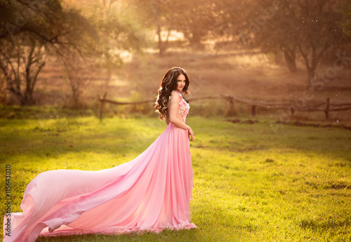 The girl in transparent pink dress with a long train flying, fairytale princess stands on top of the green hills, creative computer color fashionable toning