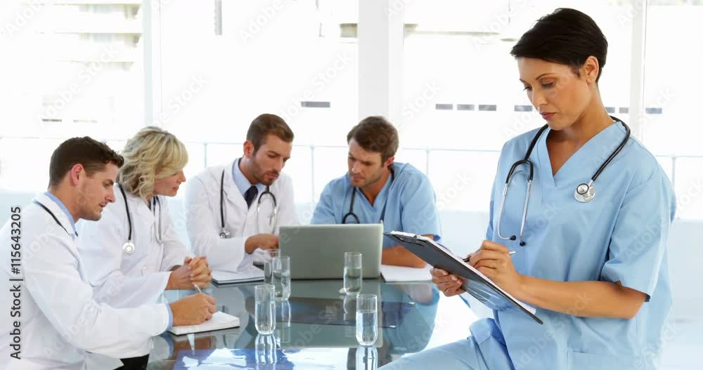 Thoughtful surgeon writing on clipboard with staff talking behind her Thoughtful surgeon writing on clipboard with staff talking behind her